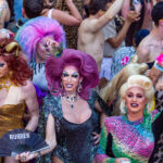 A pride parade featuring some drag queens with very brightly colored hair and clothing in the foreground amidst a general crowd including several shirtless men in the background.