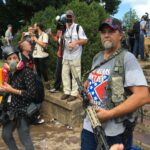 Crowd of people in a mark, nearest the camera is a middle-aged white man holding what appears to be a high-powered rifle of some sort, wearing a t-shirt festooned with the Confederate Flag and the partially visible caption "these colors don't run."