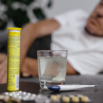 Cold medicine on a black, shiny nightstand surface in the close foreground, with an older man out of focus in the background, lying on what appears to be a couch.