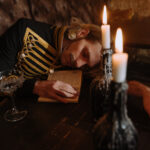 A man with unkempt shoulder-length hair is asleep on his own arm at the table of what appears to be a 19-century inn or tavern. He's wearing a formal military type of jacket common to the colonial and very early industrial periods. There is a glass cup ("crystal goblet," to be sure) in front of him and two bottle-candles sit atop the table between the man and the viewer